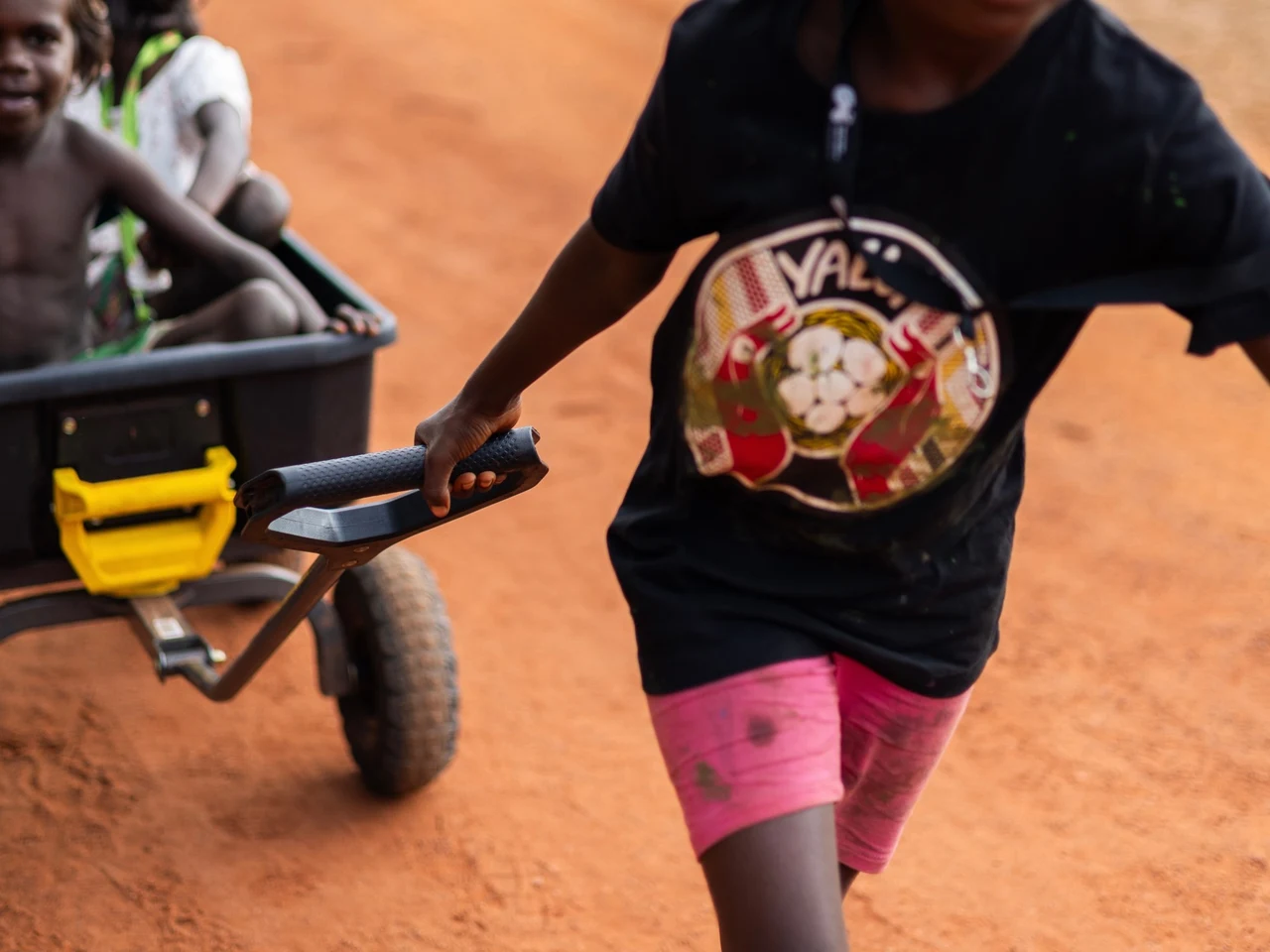 A child pulling two others in a cart along a red dirt road.