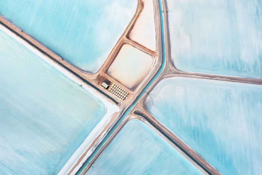 Salt ponds seen from above, pale blue fields divided by red earth roads.